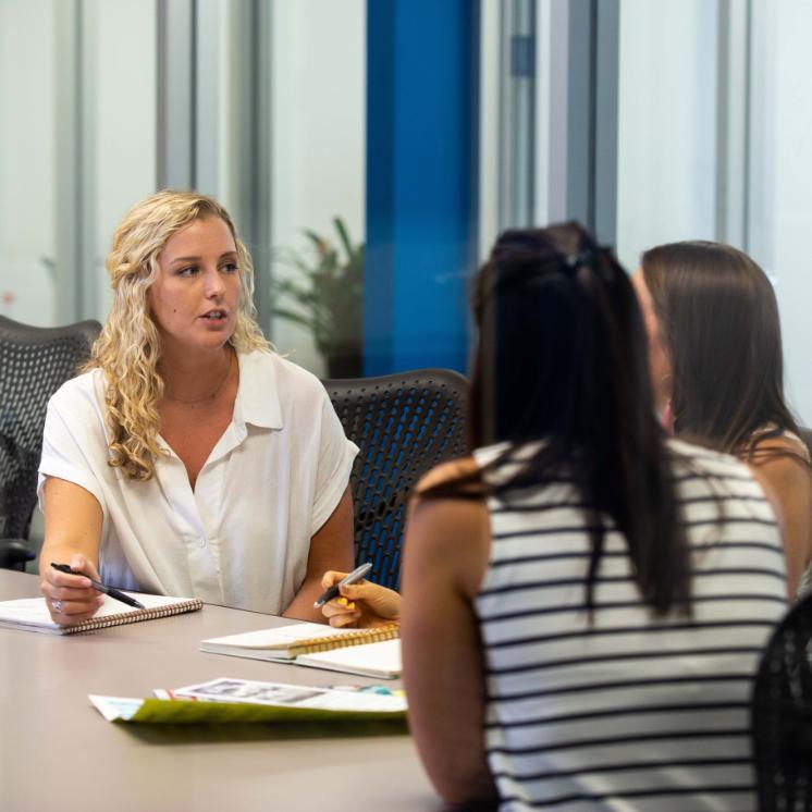 Business student during internship - Three women sitting around a conference table in discussion. One woman with curly blonde hair is speaking while holding a pen. Notebooks and papers are spread on the table. Bright modern meeting room with glass walls and natural light.
