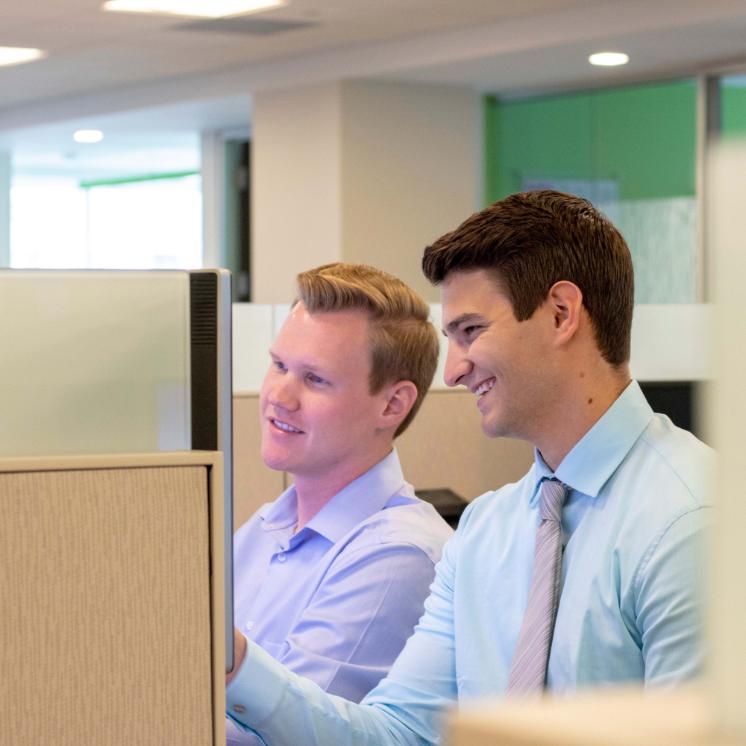 Business students working during internship - Two men sitting at office cubicles looking at a computer screen. Both are smiling and wearing dress shirts, one with a tie. They appear to be collaborating on a project. Bright modern office with glass partitions and soft lighting.