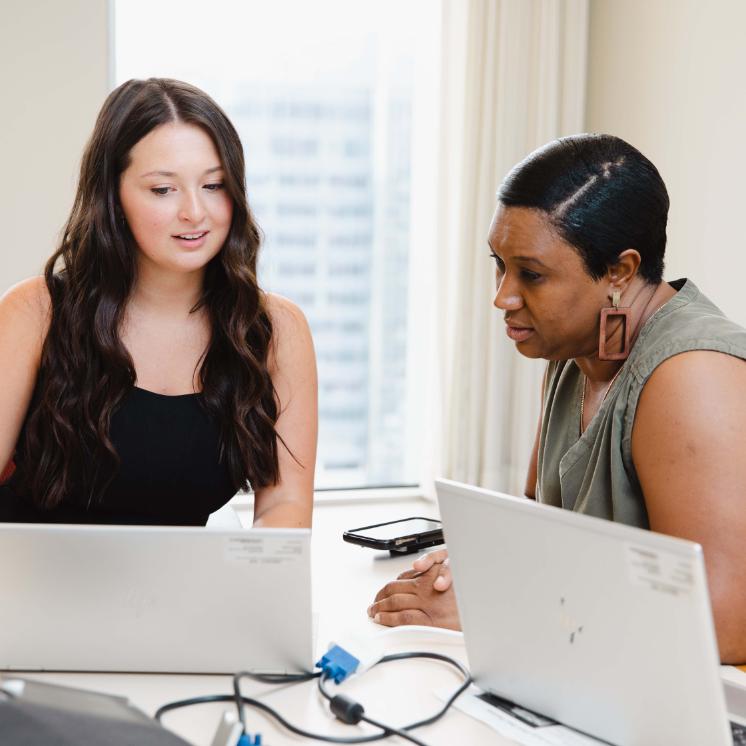 Charlotte Austin Interning at the Fed - Two women sitting at a desk with open laptops. They are focused on the screen and engaged in discussion. One has long brown hair, and the other has short dark hair. Bright office setting with window and city buildings in background.