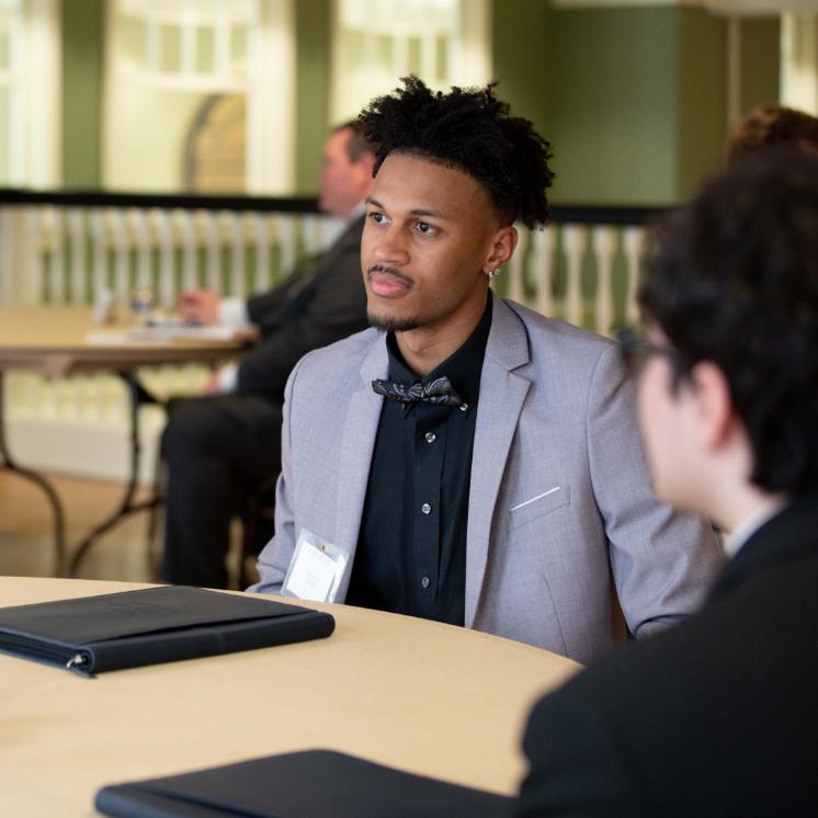 Business student participating in mock interview - Man wearing a light gray suit jacket and black shirt with a bow tie. He is seated at a round table with a folder in front of him. Other people are visible in the blurred background. Indoor professional setting with soft lighting and green walls.