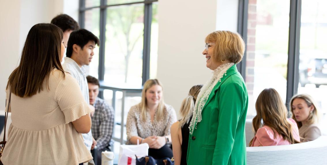 Patti Carey connecting with students at a business school event - A woman in a bright green blazer smiles while speaking with a group of students. They are gathered in a modern, sunlit space with large windows and light-colored furniture. Some students sit chatting in the background while others stand nearby. The atmosphere feels friendly and conversational.