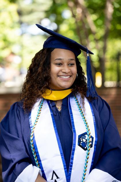 A student stands wearing academic regalia and smiles looking out of the frame. 