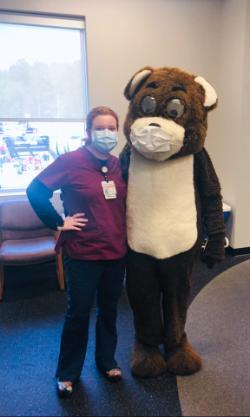 Macey Mills ’19 in scrubs at work masked standing with a mascot