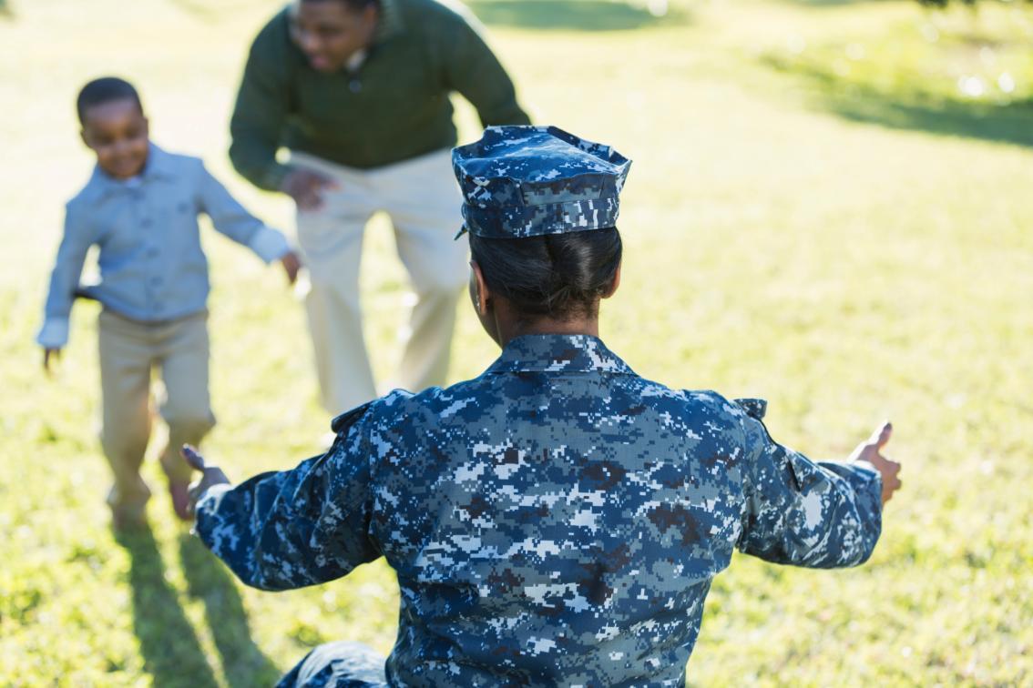 A military homecoming. rear view of an African American woman wearing a navy camouflage uniform, arms outstretched ready to welcome her little boy who is running toward her with his father.
