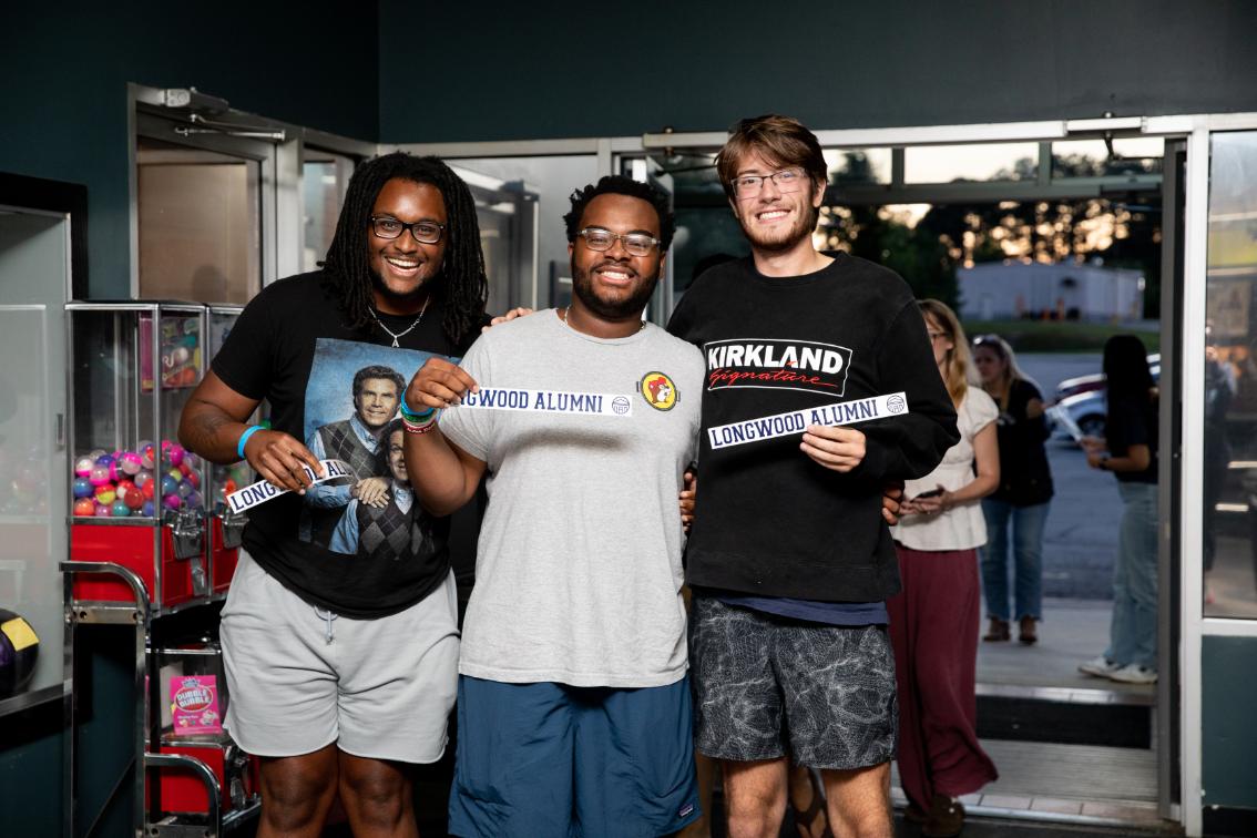 Three Longwood seniors smile together at a Senior Week bowling event while holding Longwood Alumni signs.