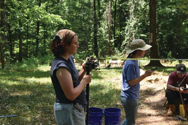 Students conduct fieldwork in a wooded area, with one holding a camera rig while another measures distance with a tape stretched between them, as a third person crouches nearby checking a device, and buckets and tools are arranged on the ground around them.
