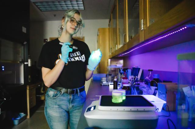 A student wearing gloves and a black T-shirt with the words “I am enough” works in a dimly lit science lab, holding a flashlight over a glowing green sample. The lab bench is lined with equipment, bottles, and instruments, illuminated by purple LED lights.