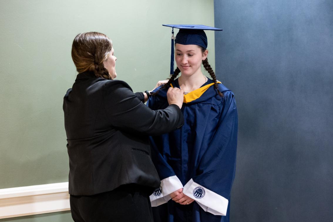 A Longwood senior wears commencement regalia as a staff member helps with the finishing touches at 100 Days to the Tassel Turns.