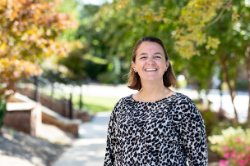 A smiling woman with straight, dark blonde hair stands outdoors in a long-sleeve top with a black-and-white leopard print pattern. Bright greenery and pink flowers appear in the background.