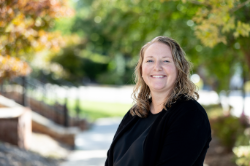 A smiling woman with shoulder-length, curly light brown hair stands outside. She wears a black top and black cardigan. The background is softly blurred with green foliage and pink flowers.