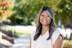 A smiling woman with long, wavy dark hair featuring blonde highlights stands outdoors. She wears a sleeveless cream sweater and a silver heart necklace. The background shows a sunny, tree-lined path with fall foliage.