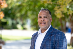 A smiling man in a white shirt and navy plaid blazer stands outdoors. He has short-cropped black and gray hair and a trimmed beard. Trees with green and yellow leaves create a softly blurred background.