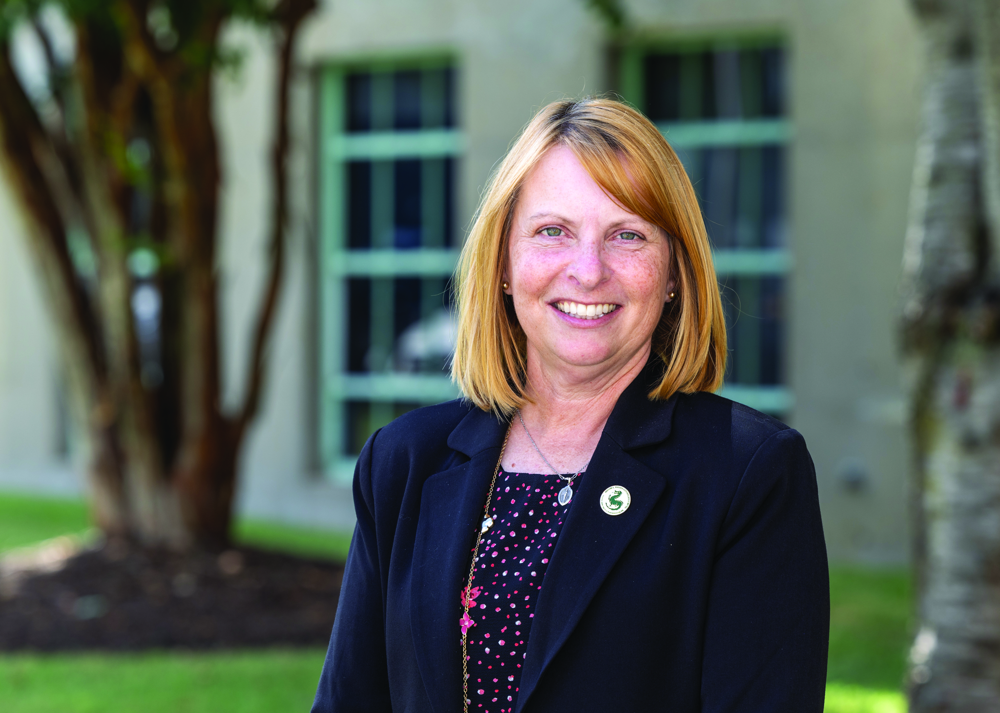 A woman with shoulder-length blonde hair smiles at the camera outdoors. She wears a black blazer over a dark patterned blouse and a necklace. A small circular pin is attached to her lapel. Blurred campus buildings and trees appear in the background.