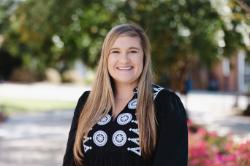 A woman with long blond hair smiles outdoors, wearing a black top with white embroidered designs, with green trees and pink flowers blurred behind her.