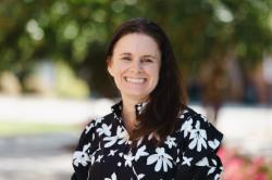 A woman smiles broadly outdoors, wearing a black blouse with large white floral patterns, with sunlight and greenery behind her.