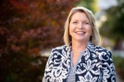 A woman with blond hair smiles outdoors, wearing a black-and-white patterned jacket and gold jewelry, with fall foliage in the background.