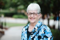 An older woman with short white hair and glasses smiles outdoors, wearing a blue patterned jacket and pearl necklace, with greenery in the background.
