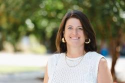 A woman smiles outdoors in a sleeveless white knit top with gold hoop earrings and layered necklaces, standing against a sunlit background of trees.
