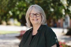A woman with gray hair and glasses smiles outdoors, wearing a dark green blouse. The background is softly blurred with trees and sunlight filtering through the leaves.