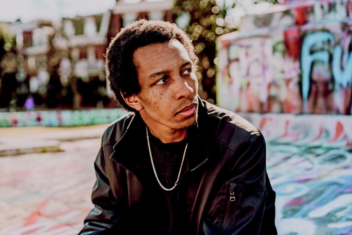 Joel Worford '18, credit: Maggie Graff/Ignited Creative 46 Man wearing a dark jacket and chain necklace sits outdoors near a graffiti-covered wall, looking off to the side with a thoughtful expression. Sunlight highlights his face, and blurred buildings and trees are visible in the background.