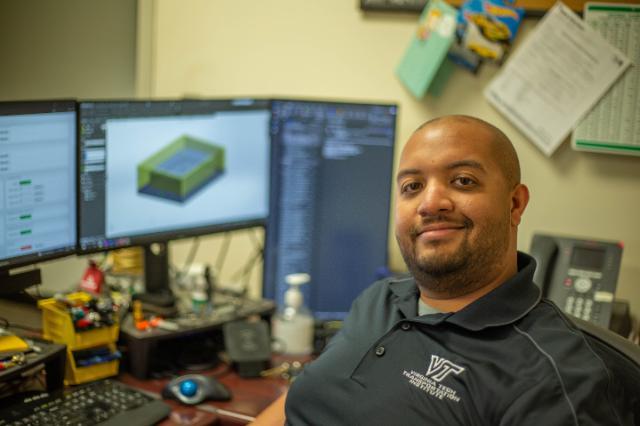 Courtesy of Virginia Tech Transportation Institute Man seated at a desk with dual monitors displaying design software, wearing a navy polo shirt embroidered with 