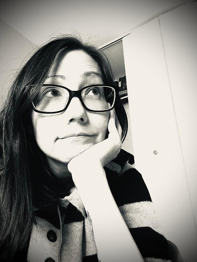 Black-and-white photo of a woman with long dark hair wearing glasses and a striped sweater, resting her chin on her hand and gazing upward thoughtfully. A closet and ceiling corner are visible in the background.