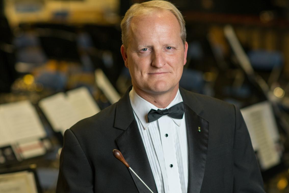 Man in a tuxedo with a bow tie holding a conductor’s baton, standing in front of blurred music stands and chairs.