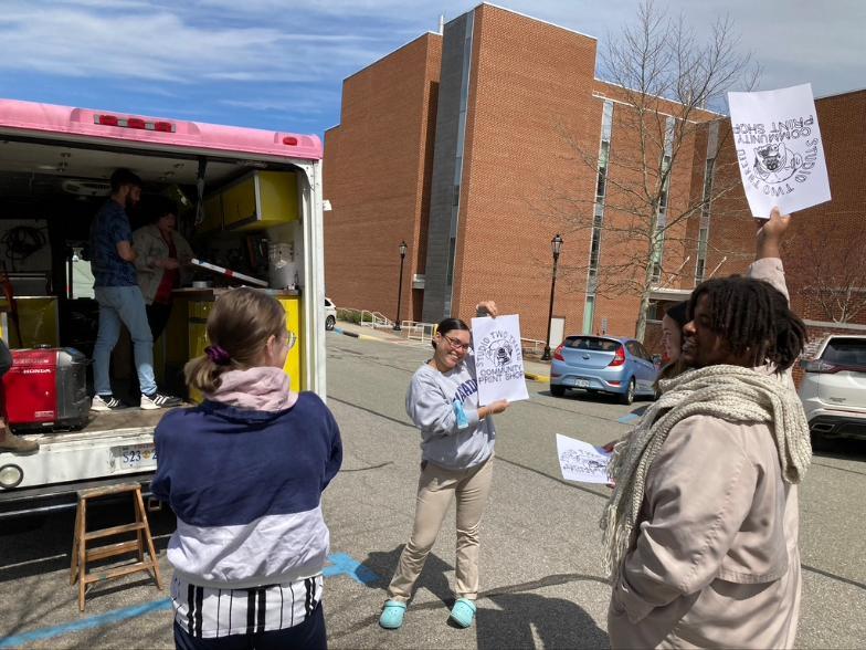 People stand outside near an open art truck with a yellow interior, smiling and holding up freshly printed posters that read “Studio Two Three Community Print Shop.” Two individuals are visible inside the truck working with screen-printing equipment, while others outside show off their prints in the sun with a brick building and parked cars in the background.