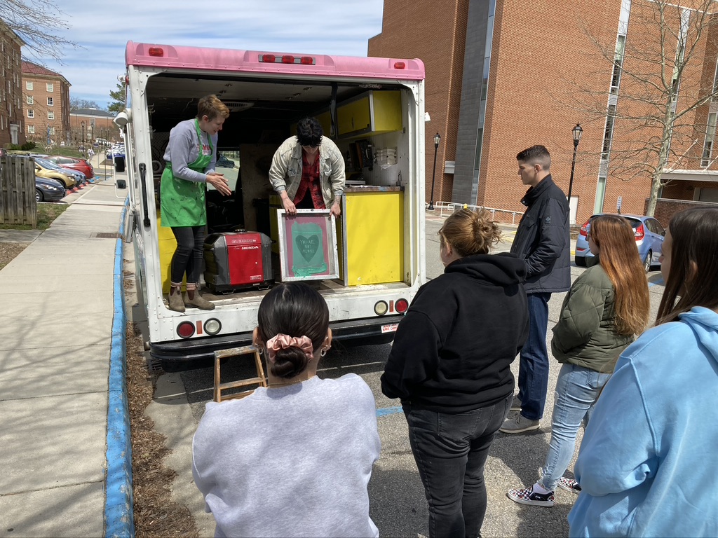 A group of people stands outside behind a parked art truck with its back doors open. Two individuals inside the truck demonstrate screen-printing equipment—one holding up a green screen frame with a heart design. The truck’s interior is painted yellow, and supplies are visible inside while participants watch from the sidewalk and street.