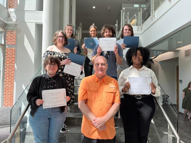 A group of eight people pose on a staircase inside a bright, modern building. Seven individuals—primarily students—stand on the steps holding award certificates and folders, while an older man in an orange shirt stands at the front center smiling. The certificates reference scholarships and awards such as “The Bushey Scholarship” and “Lucile Walsh Art Scholarship,” indicating an event celebrating