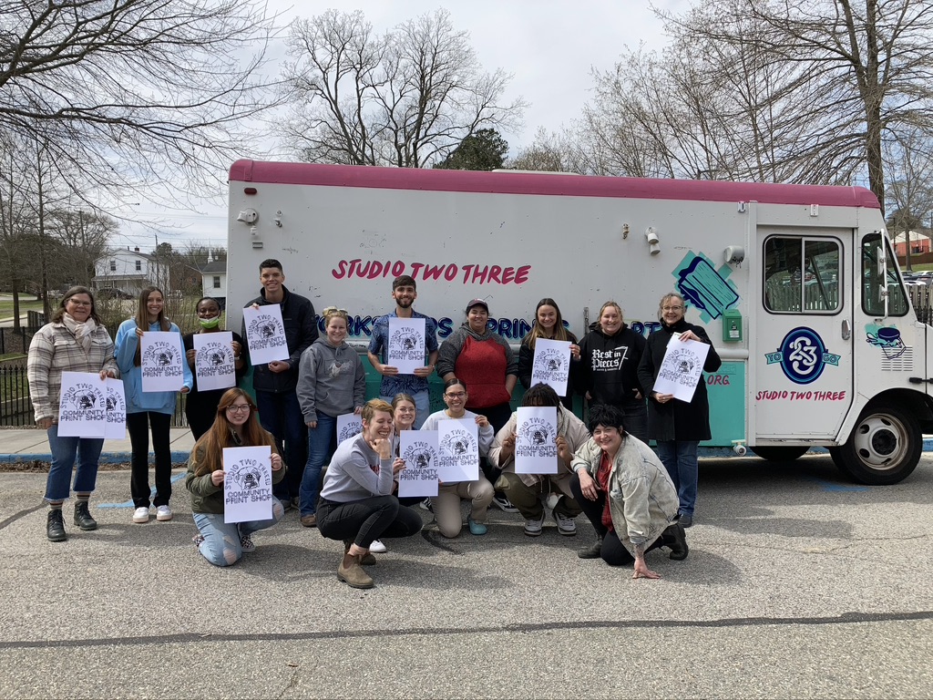 A group of people stands in front of a colorful mobile art truck labeled “Studio Two Three” holding up black-and-white printed posters that say “Community Print Shop.” The truck is painted white with teal and pink accents, featuring an illustration of a book and art supplies. Trees and houses are visible in the background on a cloudy day.