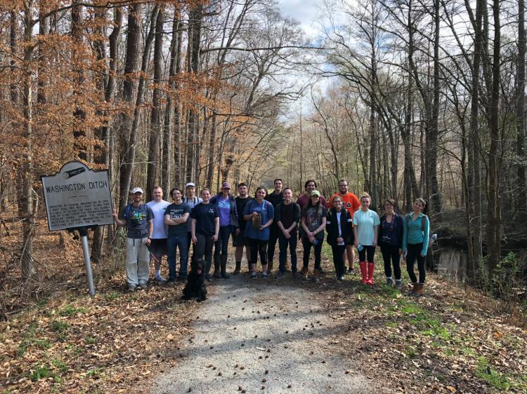 BIOL 455 students pose in front of Washington's Ditch at the Great Dismal Swamp - Group of about 18 people and a black dog standing on a wooded trail next to a historical marker sign for Washington Ditch, surrounded by tall bare and brown-leaved trees with a narrow waterway on the right.