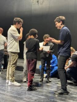 Students stand in a small circle onstage during a theater workshop or rehearsal activity.