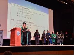 Speakers and faculty in academic regalia stand onstage during a Longwood ceremony with a schedule projected behind them.