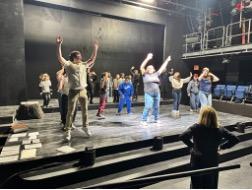 Students rehearse choreography on a black-box theater stage as a person seated in the audience area watches.