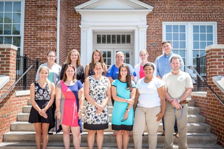14 new MBA graduates - Group of thirteen people posing on brick building steps in bright sunlight. They stand in two rows, smiling at the camera. The group includes men and women in summer business casual attire. A white doorway with columns and the number 200 is visible behind them.