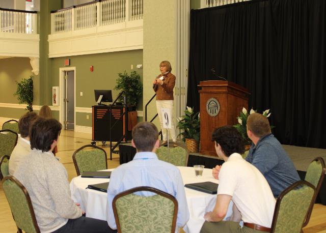 Career Cafe 5 - A woman holding a microphone speaks to students seated at round tables during a professional event in a large hall with green walls, wood accents, and a podium featuring the Longwood University seal.