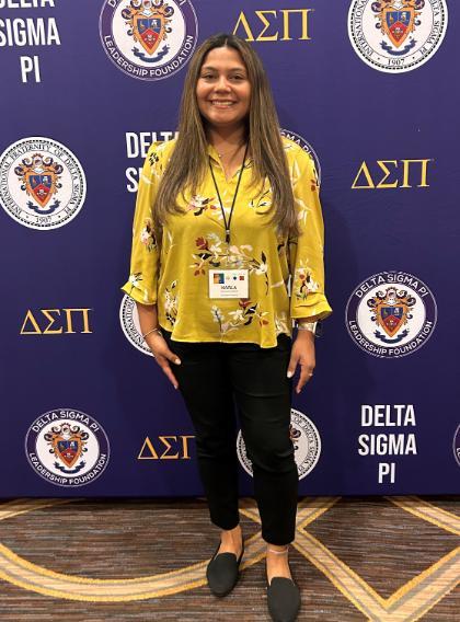 Karla Grijalva Lopez standing in front of Delta Sigma Pi step and repeat banner. - Person smiling in front of a Delta Sigma Pi backdrop. They wear a yellow floral blouse, black pants, and black shoes. A name badge hangs from a lanyard around their neck. The background features fraternity crests and Greek letters in gold.