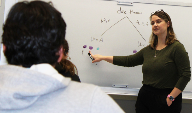 Professor standing in front of whiteboard. - Instructor teaching in front of a whiteboard with a diagram. She points to colored circles labeled under “Urn A” and “Urn B.” The chart illustrates dice outcomes split into two branches. Students sit in the foreground, viewing the lesson attentively.