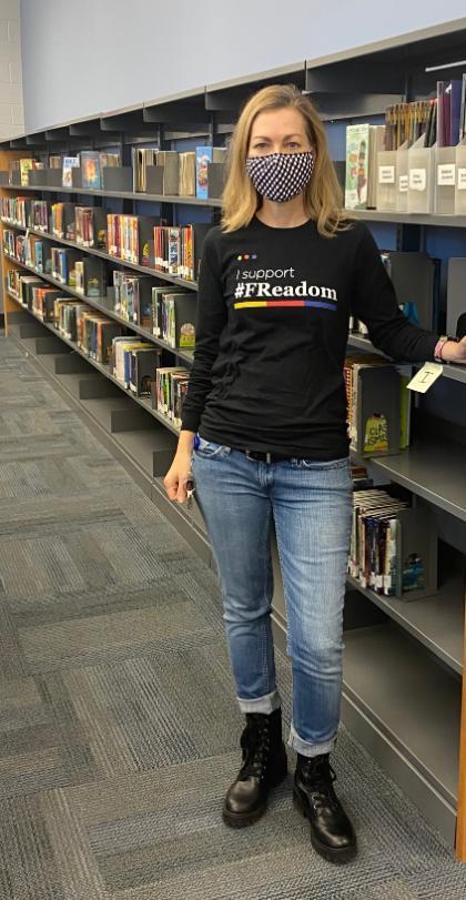 Photo of Madigan standing in a library. - A person stands in a library wearing a black “I support #FReadom” shirt, blue jeans, black boots, and a checkered face mask, with bookshelves visible in the background.