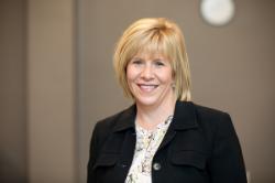 Photo of Dr. Kim Little - A person with short light-colored hair wearing a black jacket over a patterned blouse stands in front of a blurred background with neutral tones and a circular wall object.