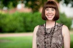 Photo of Dr. Sarah Tanner-Anderson - A person with short straight hair wearing a sleeveless patterned top and a long beaded necklace stands outdoors in a park-like setting, smiling at the camera with greenery and blurred buildings in the background.