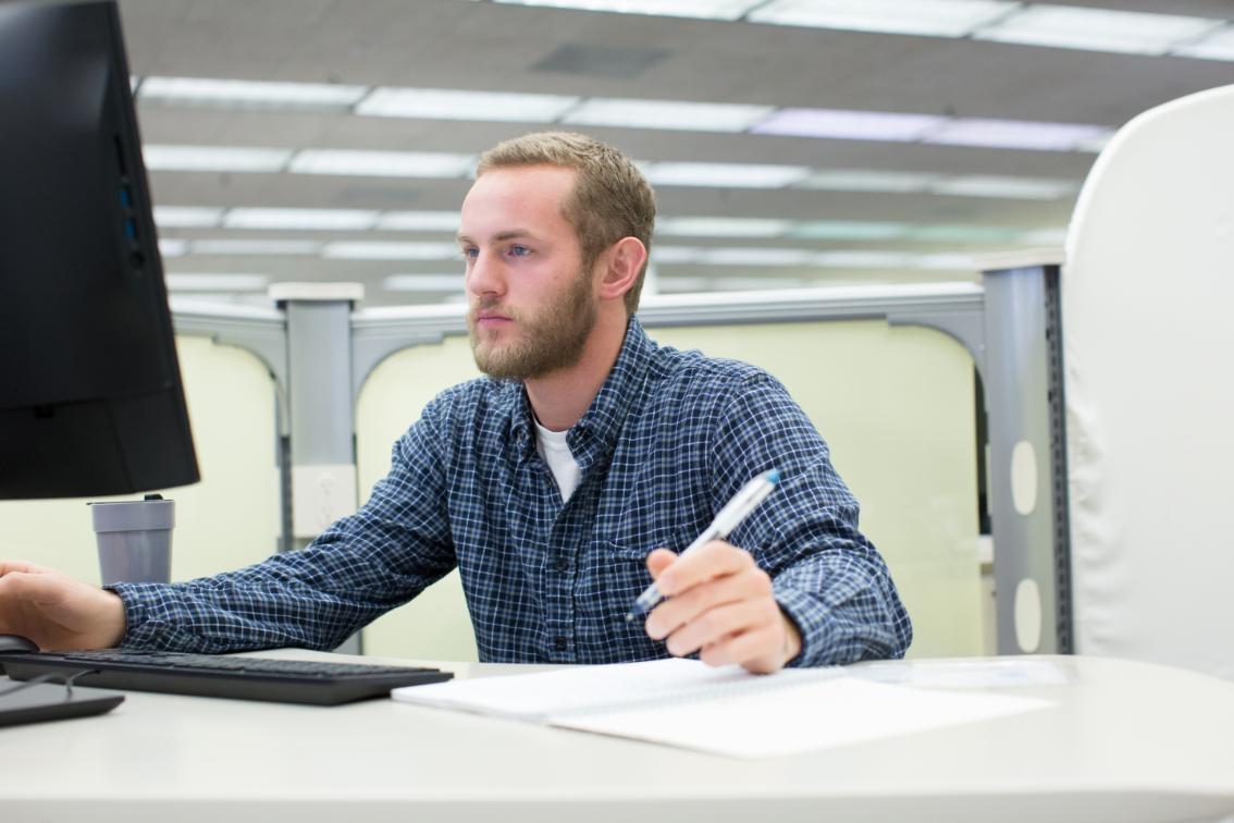 Student sits working on desktop computer - A person in a blue checkered shirt sits at a desk in an office, holding a pen and looking at a computer monitor, with a notebook, keyboard, and mouse on the desk and cubicles in the background.