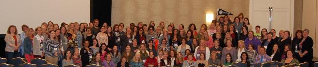 VAASL Alumni Event 2017 - A large group poses indoors in a conference or meeting room setting.   Many wear name tags, suggesting an organized event or alumni gathering.   The background includes patterned wallpaper and a wall-mounted light fixture.   One person holds a flag, indicating group or institutional affiliation.