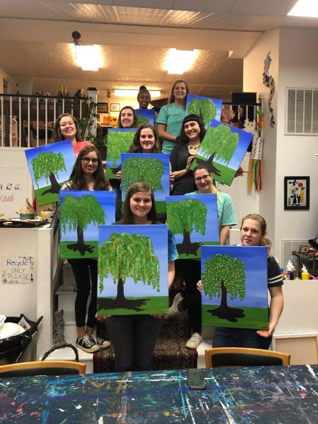 GSA Paint Night 2018 - A group of people pose in an art studio holding paintings of green trees on blue backgrounds.   They are arranged in rows, some standing on stairs, others on the ground.   The setting includes art supplies and decorations, suggesting a painting event.   Each canvas reflects a personal version of the same tree-themed artwork.
