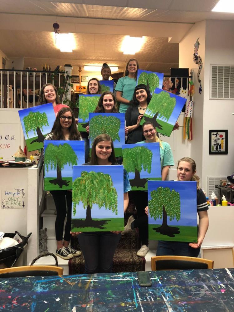 GSA Paint Night 2018 - A group of people pose in an art studio holding paintings of green trees on blue backgrounds.   They are arranged in rows, some standing on stairs, others on the ground.   The setting includes art supplies and decorations, suggesting a painting event.   Each canvas reflects a personal version of the same tree-themed artwork.