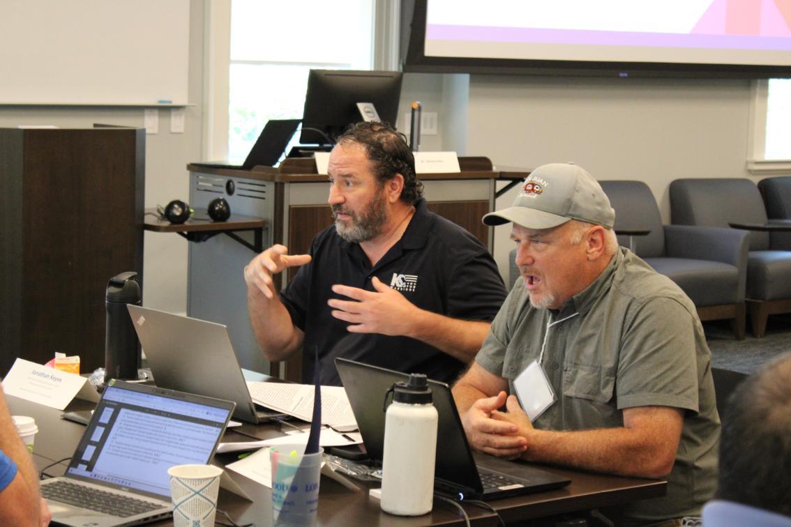 Bruce Morrow - Two people sit at a table with laptops and materials; one gestures while speaking, and the other, wearing a cap, listens attentively. Behind them are chairs, a podium, and a large screen showing part of a presentation slide.