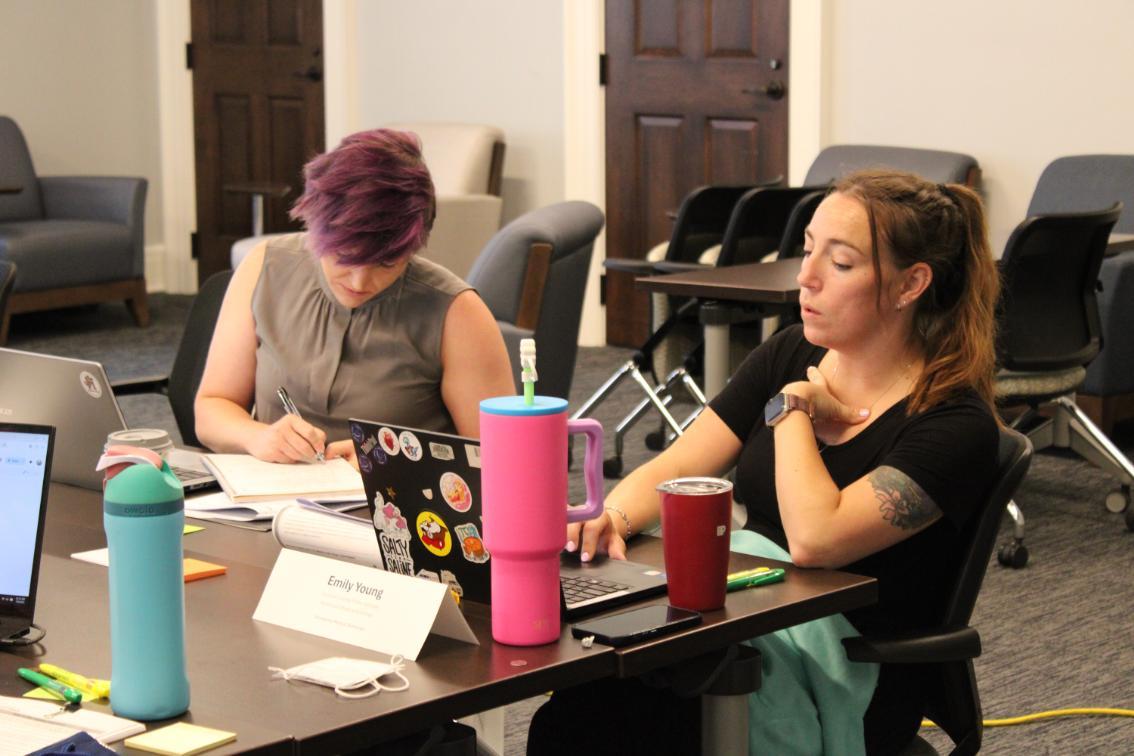 Emily Young - Two individuals sit at a table working with laptops and notebooks.   One writes in a notebook; the other focuses on a laptop screen.   Items on the table include water bottles, pens, and a nameplate reading “Emily Young.”   The setting appears to be an indoor meeting or study room.