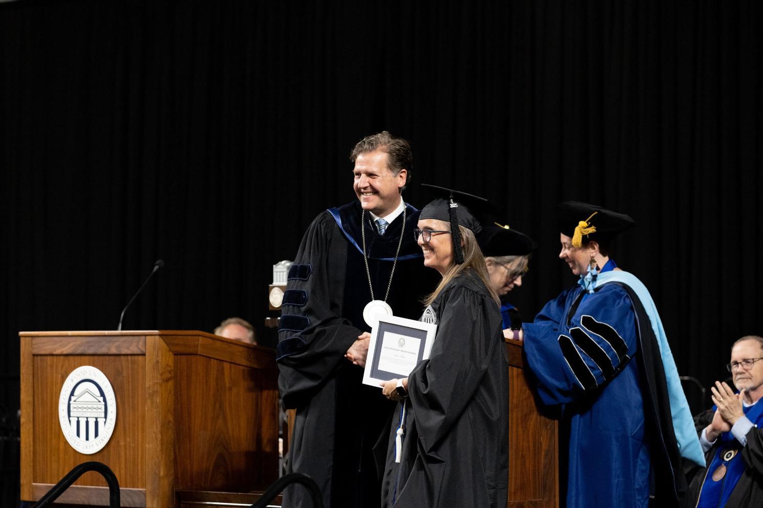 Award - A graduate in academic regalia receives a diploma on stage during commencement.   Another person in regalia presents the award while others applaud in the background.   The stage includes a podium with an emblem and formal decorations.   The scene captures a celebratory moment of academic achievement.