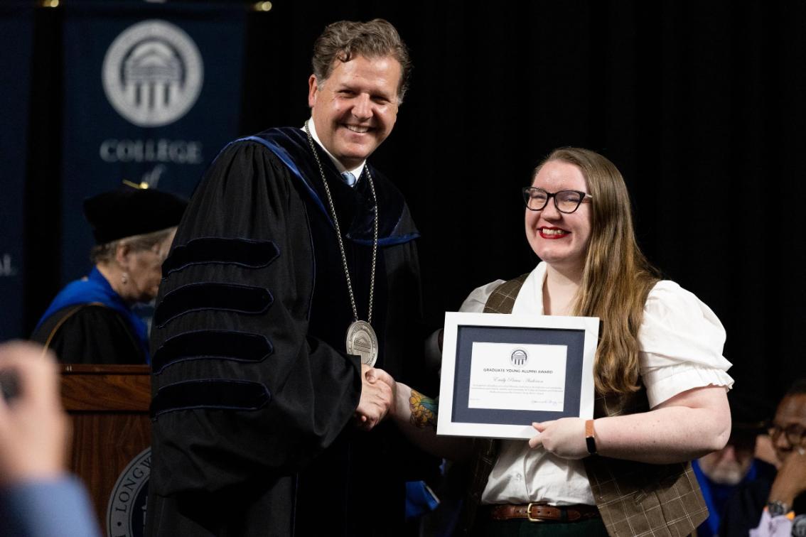 Emily Preuss Anderson - Two individuals shake hands on stage during a formal ceremony.   One wears academic regalia with a medallion; the other holds a framed certificate.   Banners with “COLLEGE” and column emblems are visible in the background.   Both are smiling, marking a moment of recognition or achievement.